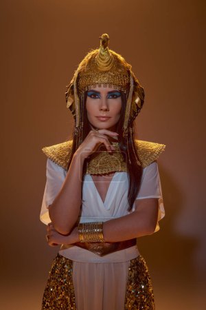 Stylish brunette woman with makeup and egyptian attire looking at camera on brown background