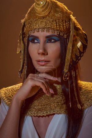 Porträt einer stilvollen Frau mit ägyptischem Kopfschmuck und kühnem Make-up, die isoliert auf braun posiert