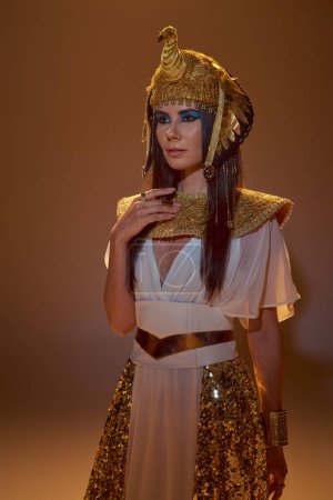 Brünette Frau in ägyptischer Kopfbedeckung und stilvollem Look posiert auf braunem Hintergrund mit Beleuchtung