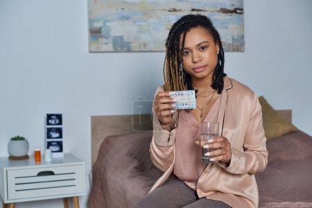 mujer afroamericana sosteniendo vaso de agua y píldoras anticonceptivas, embarazo, dormitorio moderno
