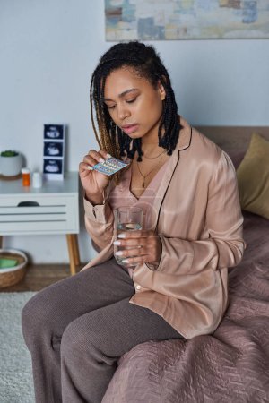 afrikanisch-amerikanische Frau hält Glas Wasser und Antibabypillen, Schwangerschaft, im Schlafzimmer sitzen