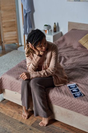 Afroamerykanka siedząca na łóżku i patrząca na zdjęcie USG, szklankę wody, antykoncepcję