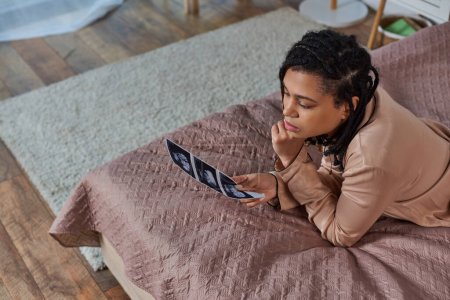 Téléchargez les photos : Vue de dessus de femme afro-américaine inquiète couchée sur le lit, regardant l'échographie, à naître, avortement - en image libre de droit