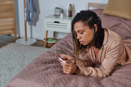 african american woman lying on bed, looking at pregnancy test, making decision, abortion concept