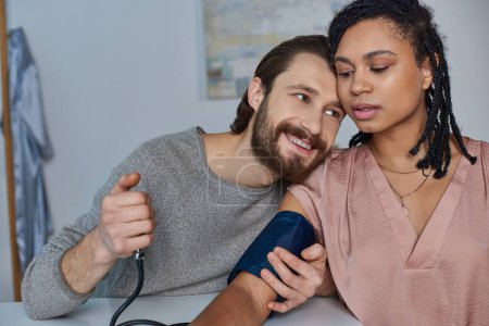 cheerful man measuring blood pressure of pregnant african american wife, future parents, expectation