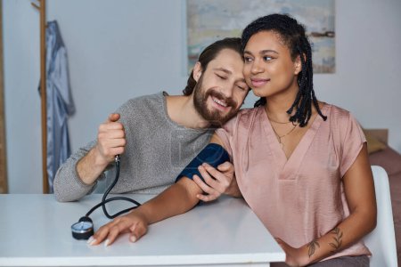 Téléchargez les photos : Homme joyeux mesurant la pression artérielle de la femme afro-américaine enceinte, futurs parents, attente - en image libre de droit