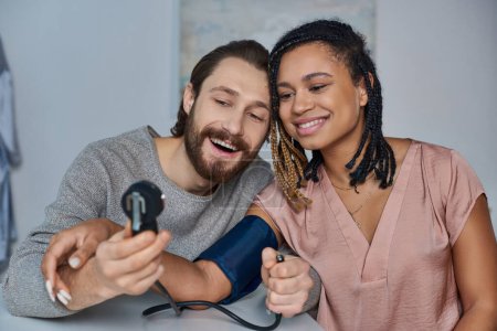 joyful man measuring blood pressure of pregnant african american wife, future parents, happiness