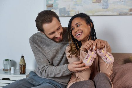 mujer afroamericana feliz sosteniendo pequeños zapatos de bebé cerca de marido, futuros padres, expectativa, lindo