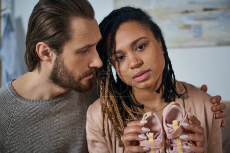 bearded man calming down african american wife crying and holding baby clothes, miscarriage concept