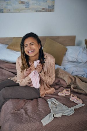 Foto de Dolor, mujer afroamericana llorando cerca de ropa de bebé en la cama, concepto de aborto espontáneo, depresión - Imagen libre de derechos