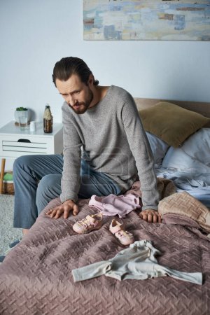 Foto de Dolor, hombre deprimido llorando cerca de la ropa de bebé, sentado en la cama, concepto de aborto espontáneo, angustia - Imagen libre de derechos