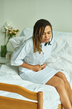 mujer afroamericana sentada en la cama, tocando el vientre, sala privada, hospital, concepto de aborto espontáneo