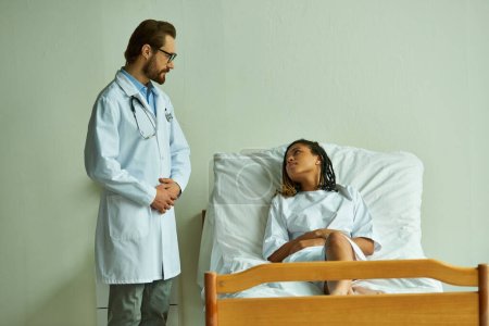 bärtige Ärztin neben afrikanisch-amerikanischer Frau im Krankenhauskleid, Privatstation, Patientin