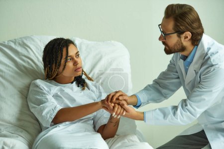 doctor holding hands of upset african american woman, comforting patient, private ward in hospital