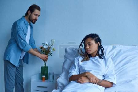 Fehlgeburtenkonzept, Mann legt Blumen in Vase neben depressiver afrikanisch-amerikanischer Frau, Krankenhausstation