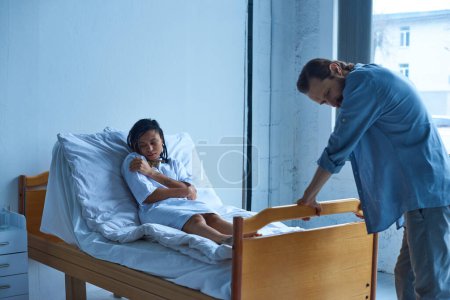 Téléchargez les photos : Concept fausse couche, femme américaine africaine déprimée couchée dans un lit d'hôpital près du mari, désespoir - en image libre de droit