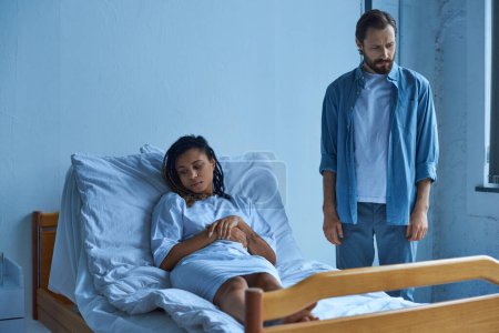 Téléchargez les photos : Concept fausse couche, triste femme afro-américaine en deuil, couché dans un lit d'hôpital près du mari, quartier - en image libre de droit