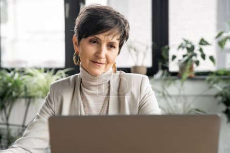 positive Geschäftsfrau mittleren Alters, die in der Nähe von Laptop im modernen Büro arbeitet, unternehmerischer Erfolg
