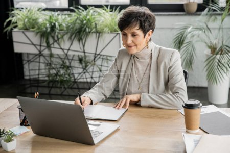 mujer de negocios experimentada escribiendo en un cuaderno cerca de la computadora portátil y bebida para llevar, planificación