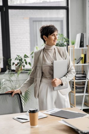 positive middle aged businesswoman with laptop looking away near documents and paper cup