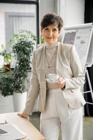 glückliche Geschäftsfrau mittleren Alters mit Kaffeetasse neben Smartphone und Dokumenten im Büro, Portrait