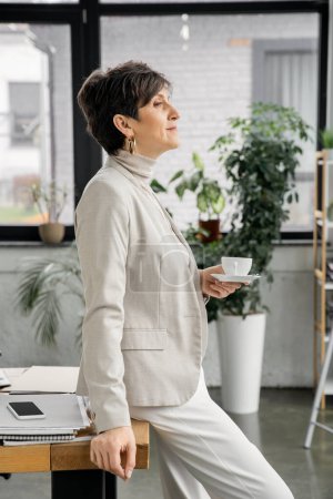 Photo for Mature businesswoman with coffee cup looking away near documents and smartphone in office, side view - Royalty Free Image