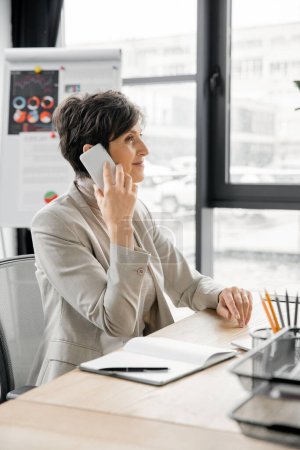 optimista gerente de mediana edad hablando en el teléfono móvil cerca de portátil en el lugar de trabajo en la oficina moderna
