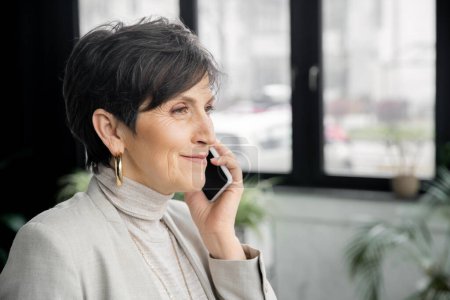 glückliche und stilvolle Geschäftsfrau mittleren Alters, die im modernen Arbeitsumfeld mit dem Handy telefoniert