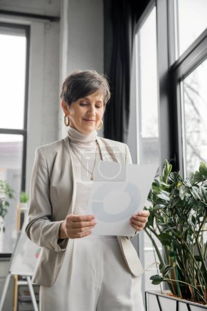 mature optimistic businesswoman looking at document with graphs in office, productivity, growth