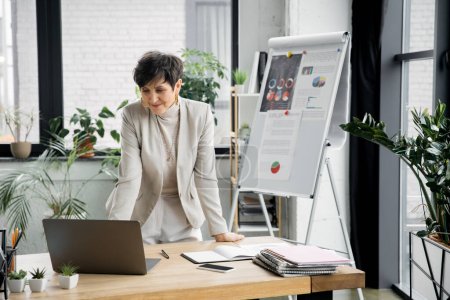 smiling middle aged businesswoman near laptop and flip chart with graphs, video call, conference
