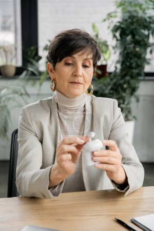 Geschäftsfrau mittleren Alters hält Kopfhörertasche mit Ohrhörern am Arbeitsplatz im modernen Büro