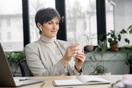 pleased mature businesswoman in earbud browsing internet on smartphone near laptop in office