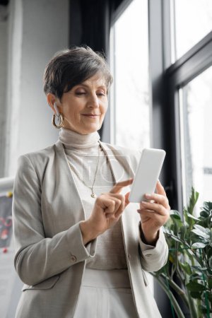 mature woman, corporate manager listening music in earbud and networking on smartphone in office