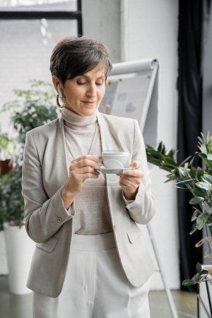 mature businesswoman holding coffee cup and listening music in earphone in modern office, enjoyment