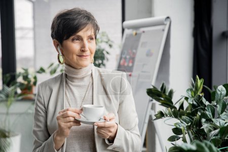 mature businesswoman with coffee cup listening music in earphone and looking away in office