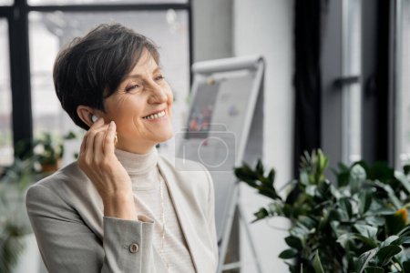 overjoyed middle aged businesswoman adjusting earphone while listening music in office, enjoyment