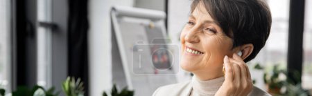 pleased middle aged entrepreneur adjusting earbud and listening music in office, banner