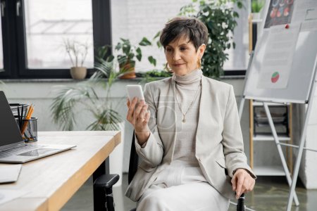 positive middle aged businesswoman with smartphone near laptop and flip chart on blurred background
