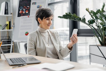 Geschäftsfrau mittleren Alters mit Smartphone neben Laptop und verschwommenem Flipchart mit Grafiken im Büro