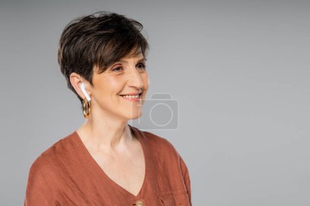 delighted middle aged woman in earbud listening music on grey, brown jacket, fall fashion, portrait