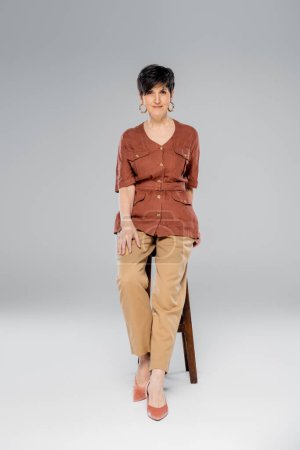 fall fashion, middle aged woman in trendy outfit sitting on high stool on grey, full length