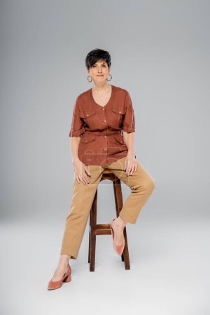 fall fashion, mature brunette woman in stylish clothes sitting on high stool on grey, full length