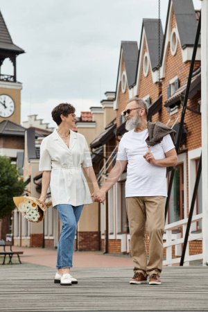 heureux couple âgé en lunettes de soleil souriant et se tenant la main, marchant ensemble, faisant du shopping, été