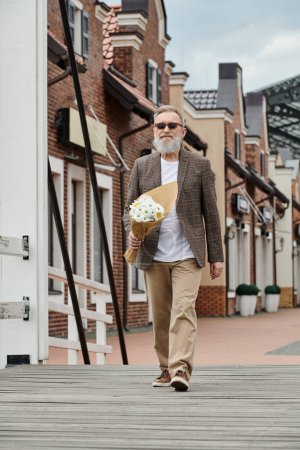 senior man with beard and sunglasses holding bouquet of flowers, walking on urban street, stylish