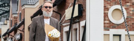 Älterer Mann mit Bart und Sonnenbrille hält Blumenstrauß in der Hand, steht auf einer städtischen Straße, Transparent