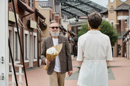 älterer Mann mit Bart und Sonnenbrille, Blumenstrauß haltend, auf der Straße auf Frau zugehend, Date, Romanze