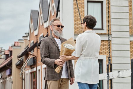 glücklicher älterer Mann mit Bart und trendiger Sonnenbrille, der Frau auf der Straße einen Strauß schenkt, Date, Romanze