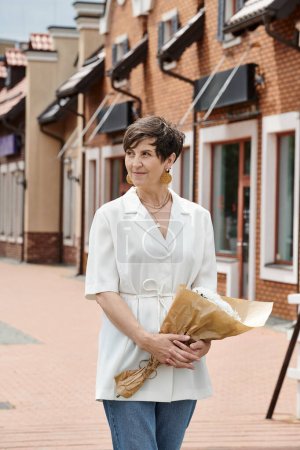 Téléchargez les photos : Femme âgée tenant bouquet de fleurs et regardant loin sur fond urbain, été, tenue blanche - en image libre de droit