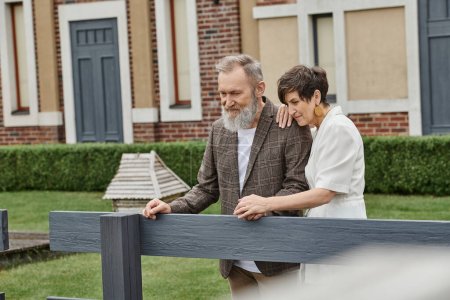 Photo for Cheerful elderly couple, man and woman standing near fence next to house and looking away, romance - Royalty Free Image