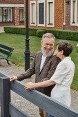 happy elderly couple, senior man and woman standing near fence next to house, looking at each other Poster #669960280